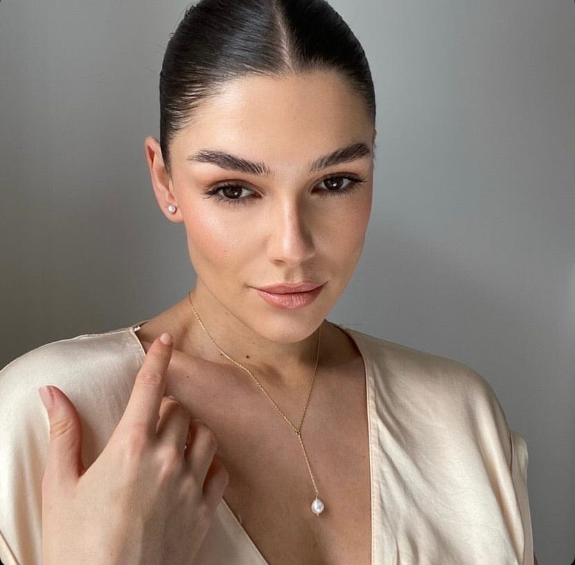 A woman with slicked-back hair wears a cream-colored top, pearl earrings, and a Bridal Pearl Drop Lariat.