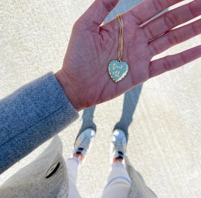 Person holding a F**K Off Necklace heart-shaped pendant with an inscription.