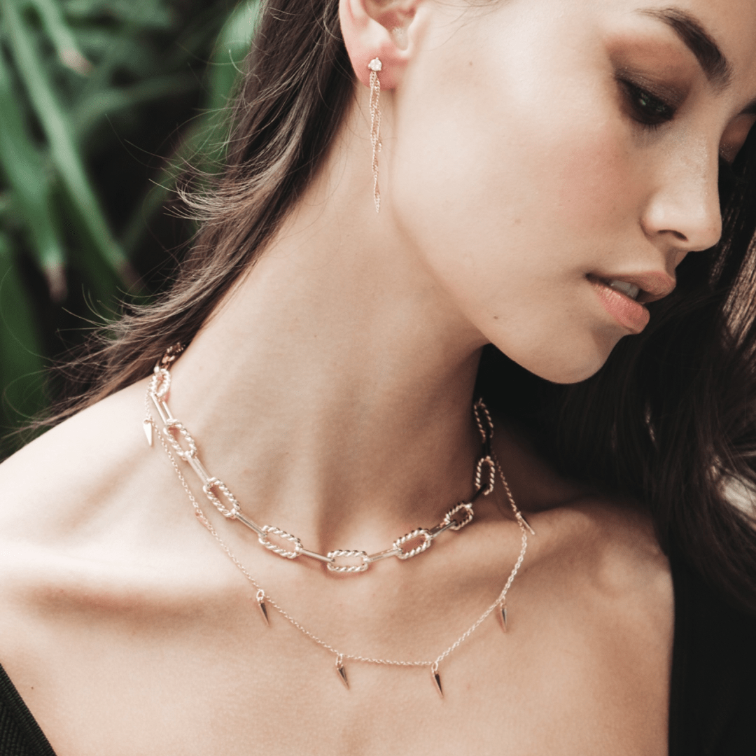 A woman wears gold Double Link Twisted Chain jewelry, including earrings and necklaces, against a leafy background.