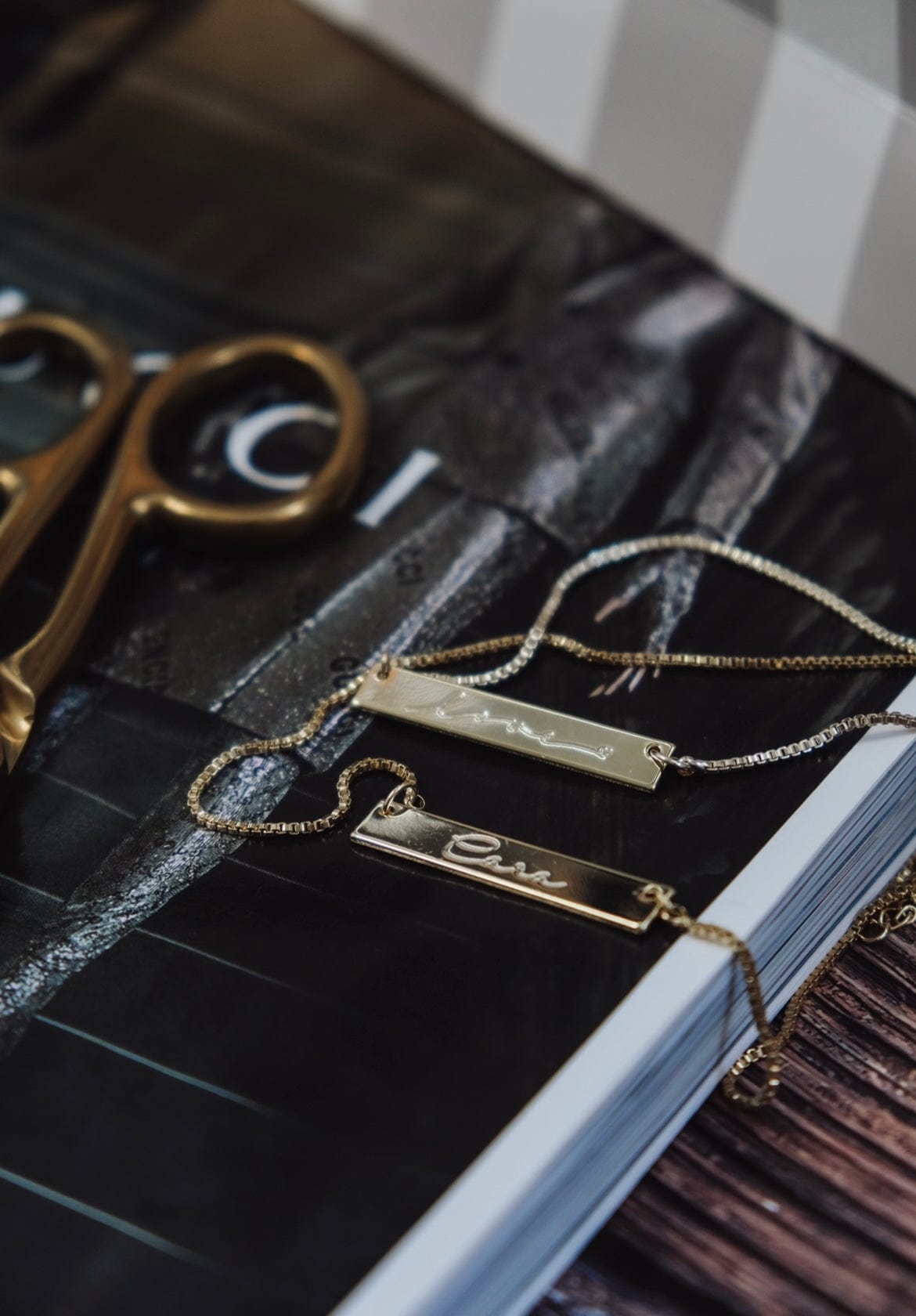 Two gold Cara Bar Necklaces with nameplates rest on a magazine next to gold scissors.