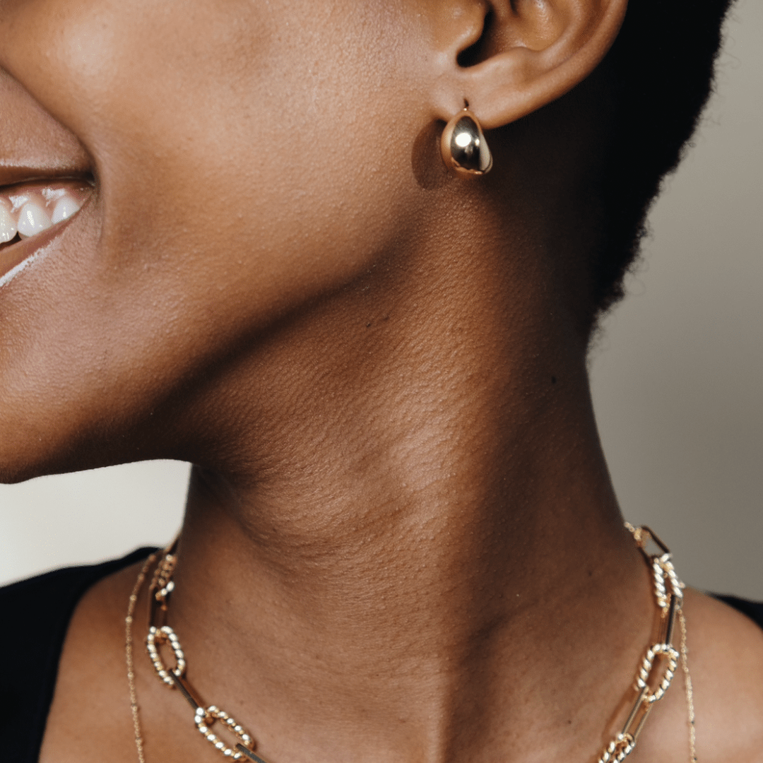 Close-up of a woman's face and neck, wearing gold Dome Hoops and a gold chain necklace, smiling slightly.