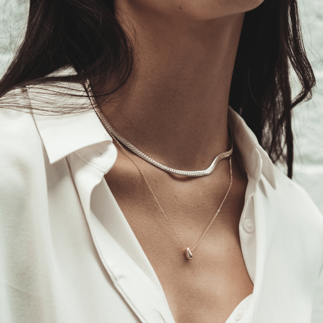 A person wears The Snake Herringbone 14 inches, a silver chain and a gold pendant necklace with a white button-down shirt.