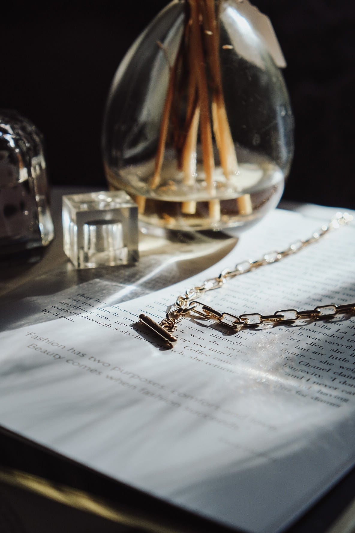 A gold T-Bar Link Chain necklace lays on an open book next to glass decorative objects.