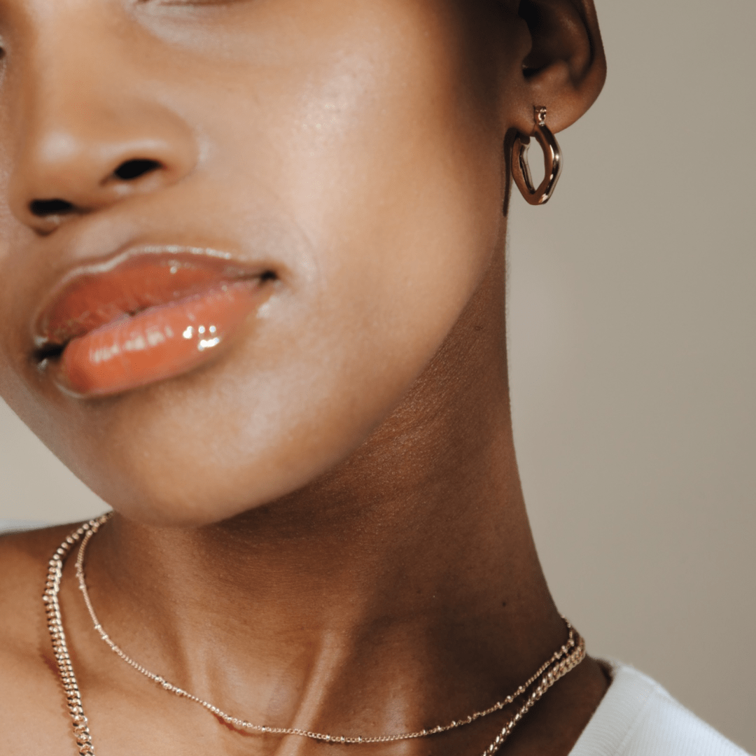 Close-up of a person's face featuring glossy lips, gold earrings, layered gold necklaces, and Chunky Melted Hoop earrings.