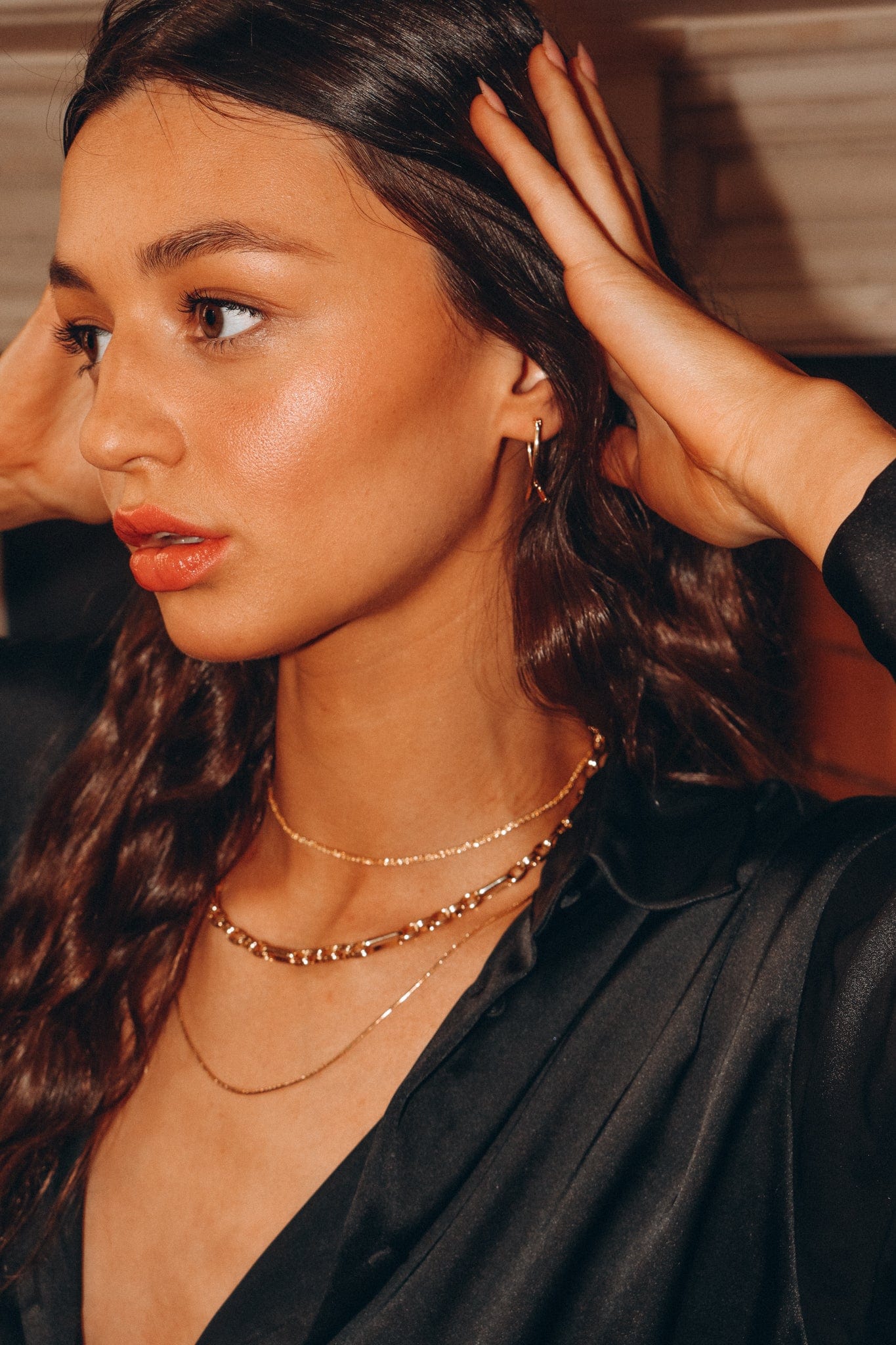 A woman wearing layered gold Box Chain 19 inches necklaces and earrings touches her hair.