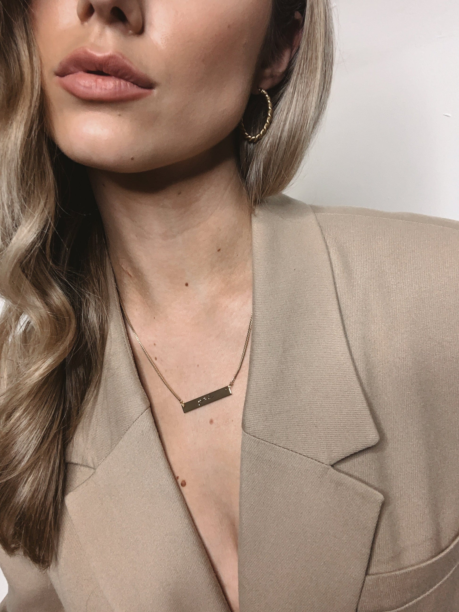 Partial view of a woman with blonde wavy hair wearing a tan blazer and a Cara Bar Necklace.