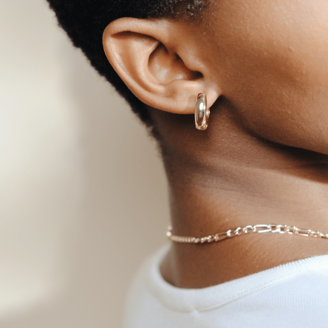 Close-up of a person's ear with a silver chunky huggie hoop earring and a gold chain necklace.