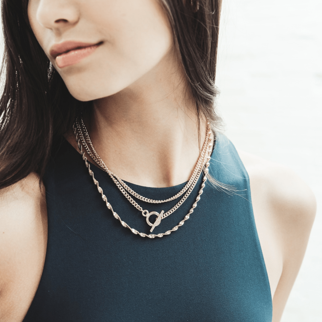 A woman wearing a blue dress and layered gold necklaces with an Inter Curb Toggle T-Bar.