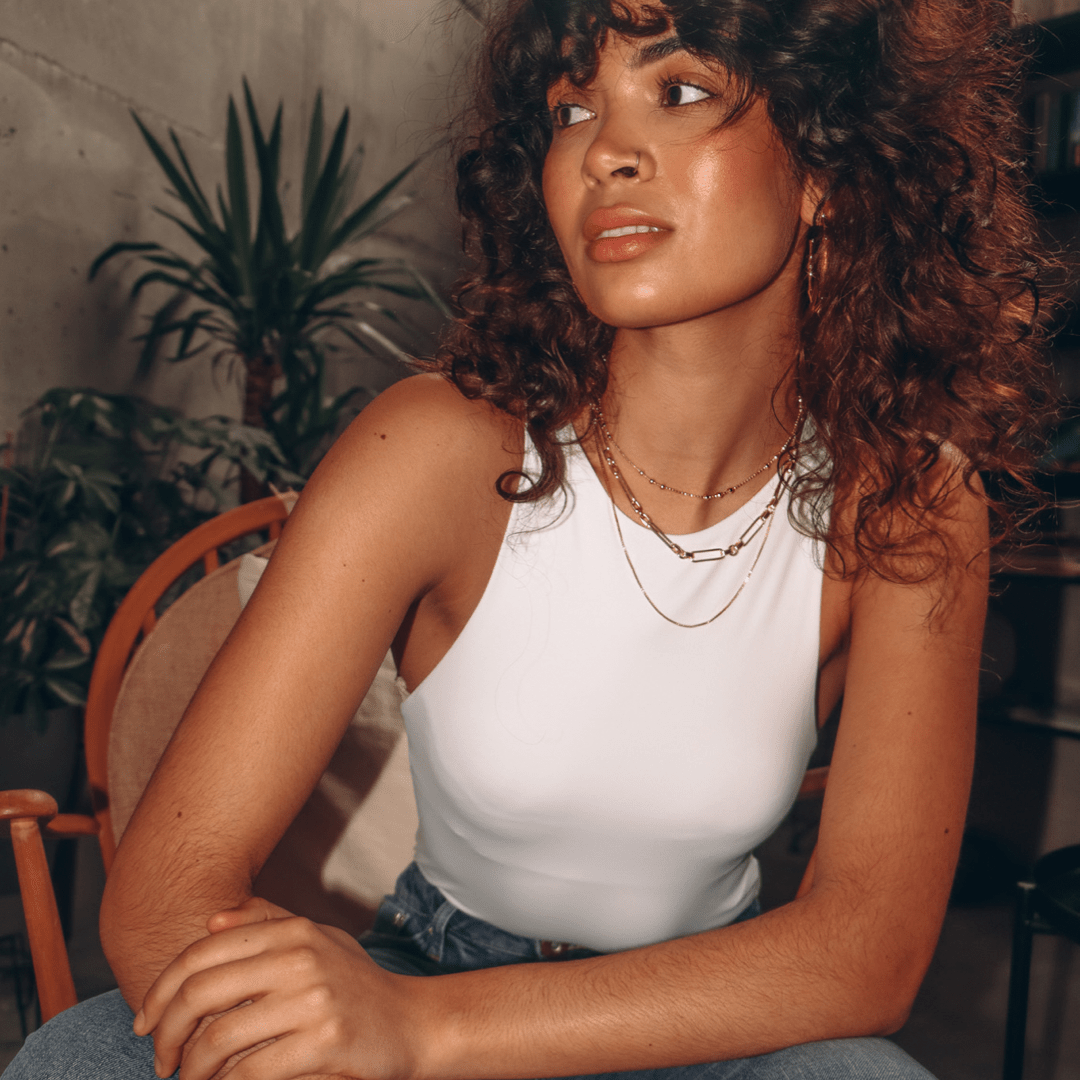 A woman with curly hair sits in a chair wearing a white tank top and layered gold Box Chain 19 inches necklaces.
