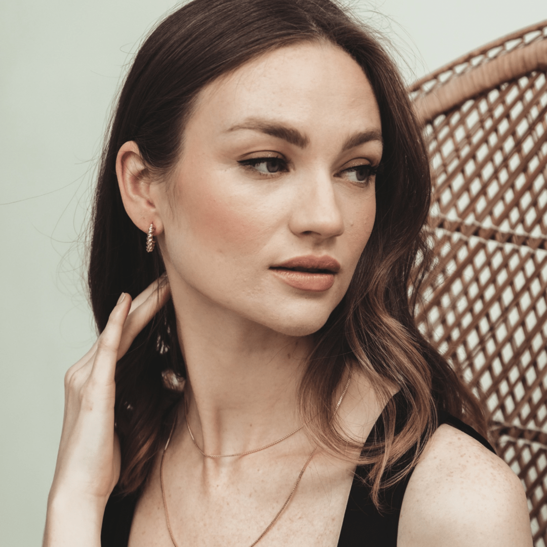 A woman with freckles and brown hair wearing Box Chain 14 inches jewelry, seated in a wicker chair, looks to the side.