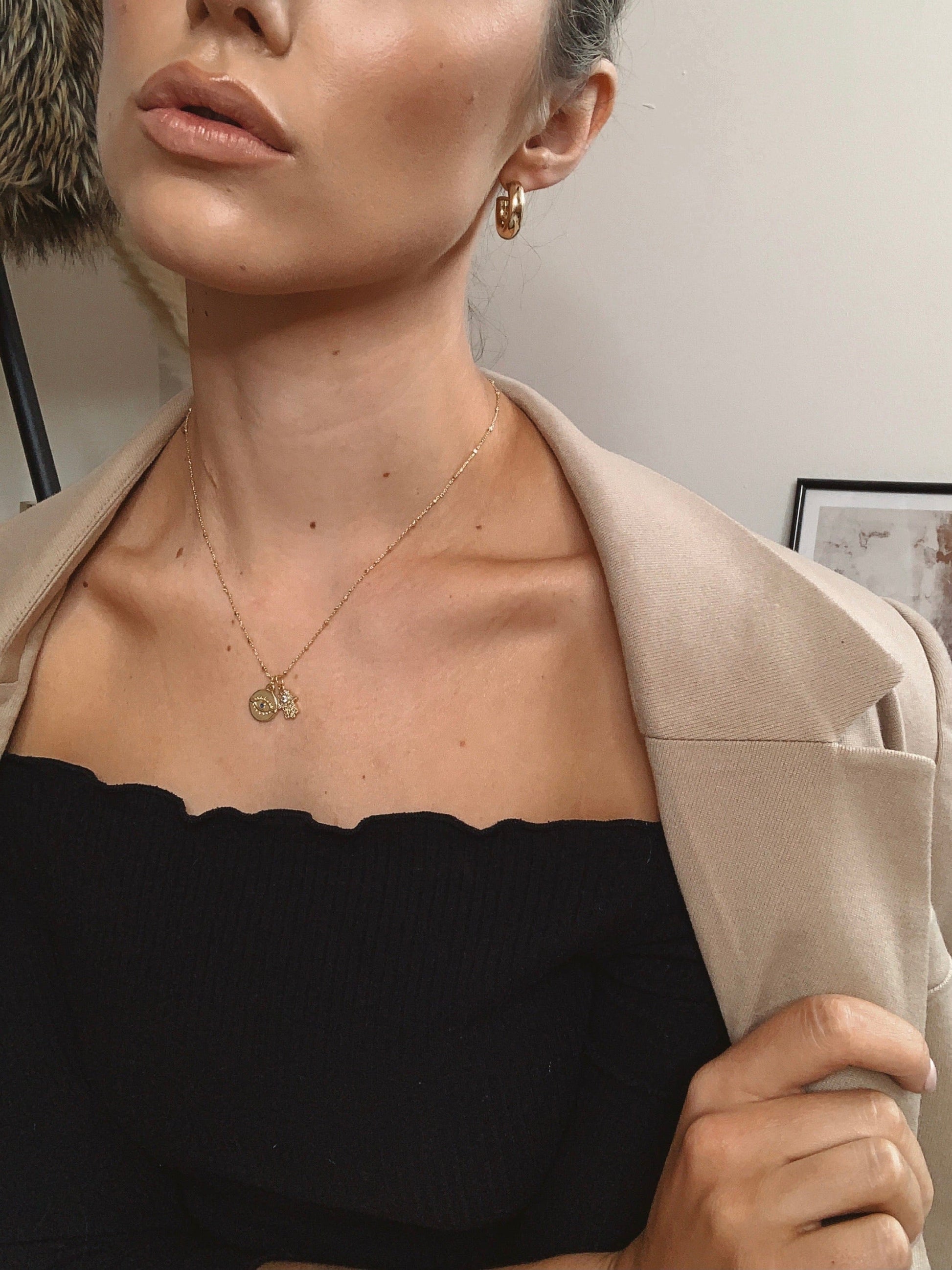 A woman wearing a tan blazer, black top, gold earrings, and a gold Evil Eye and Hamsa Hand necklace is shown in a close-up shot.