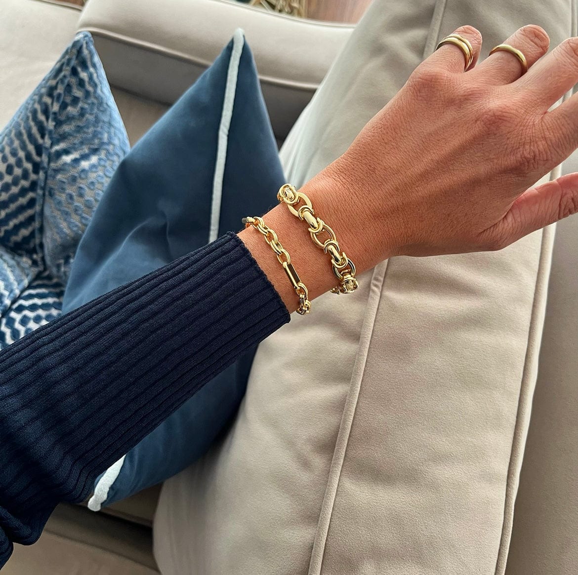 A person's hand wearing a Triple Link Bracelet and two gold rings rests on a couch.