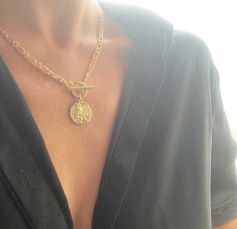 A person wearing an Athena Toggle Coin gold necklace with a round pendant, under a black shirt.