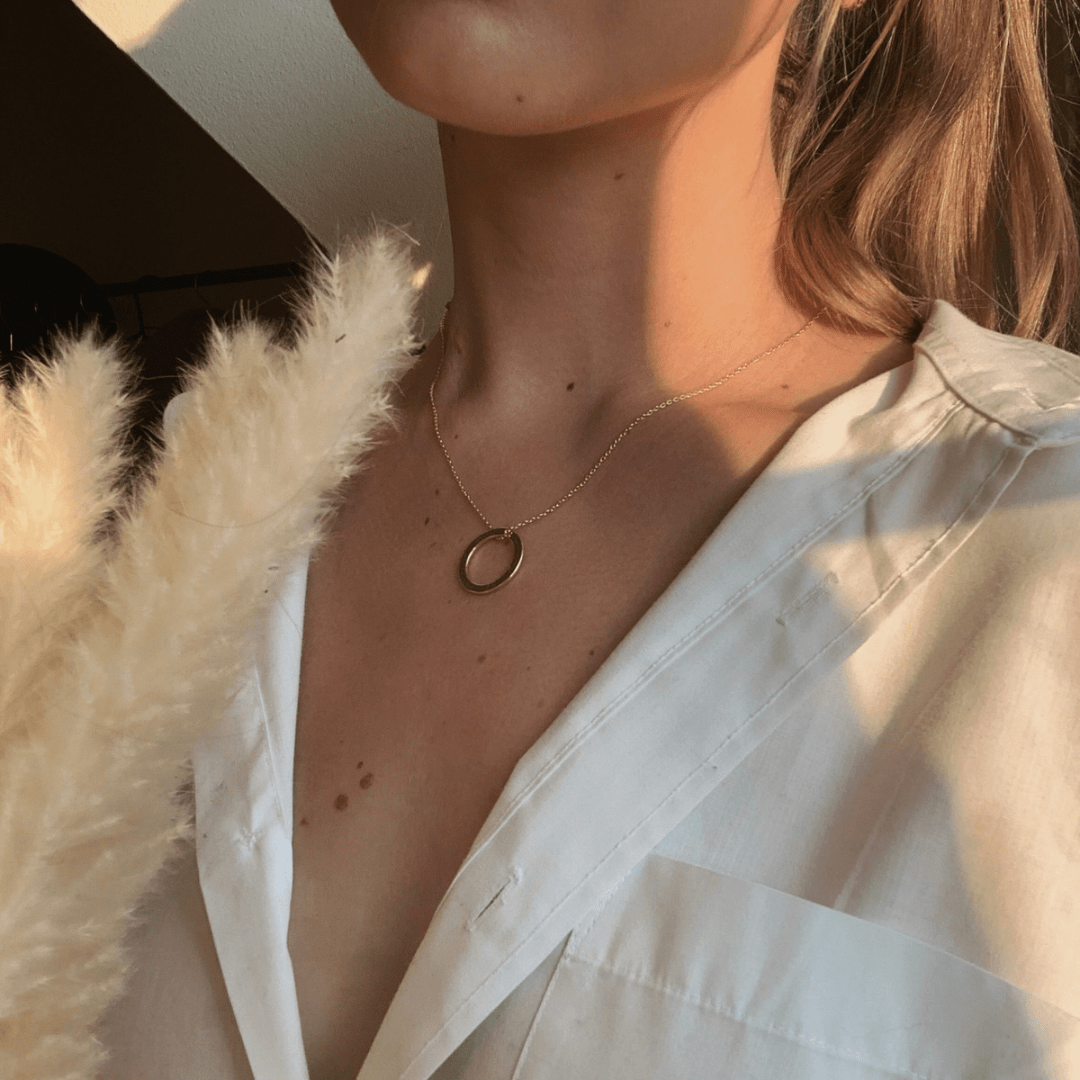 Person wearing a gold Dá fhada an lá tagann an tráthnóna necklace and a white shirt, holding pampas grass.