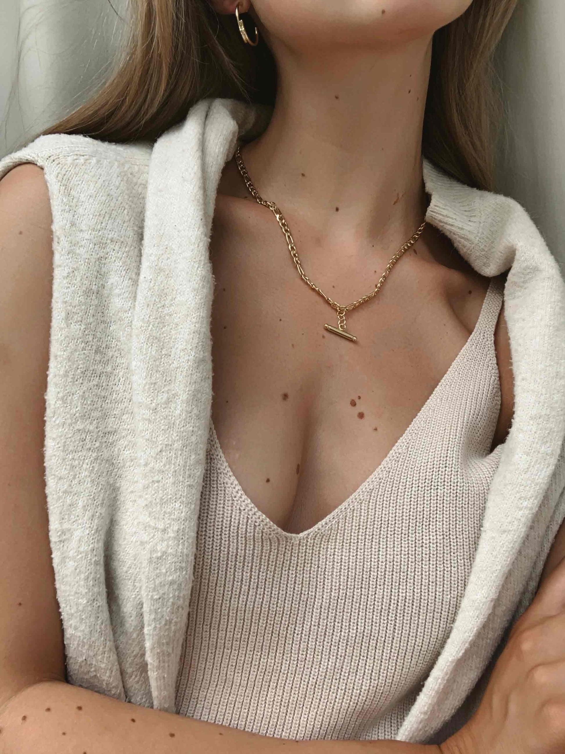 A person wearing a tank top and a vest, accessorized with a gold T-Bar Figaro Necklace and earrings.