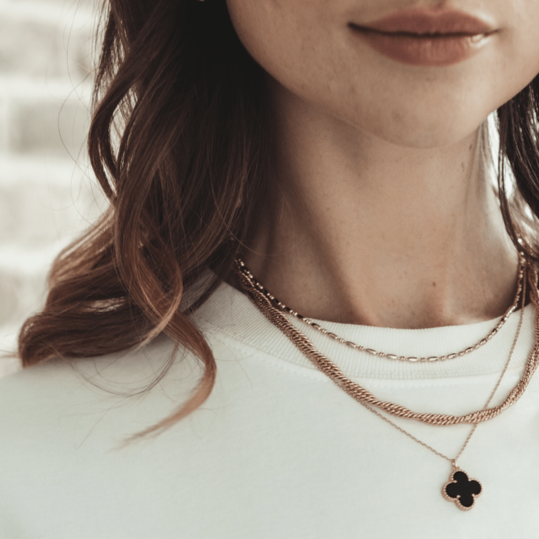 Partial view of a person wearing a white shirt and layered gold necklaces, one with a black clover pendant, including the Crossing Twisted Chain.