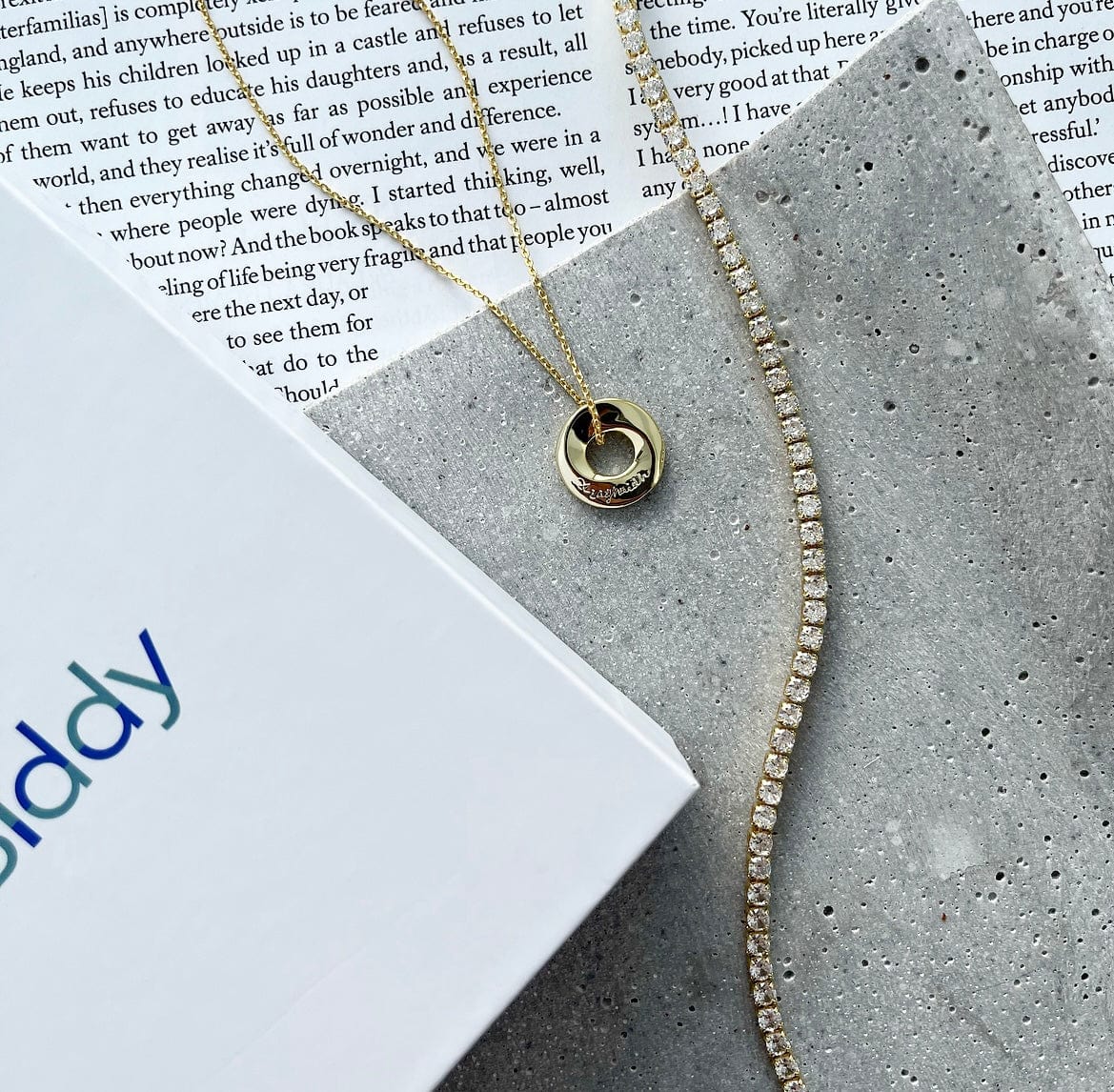 A gold Ar Aghaidh necklace and diamond tennis bracelet are displayed on a textured surface with a book and white box.