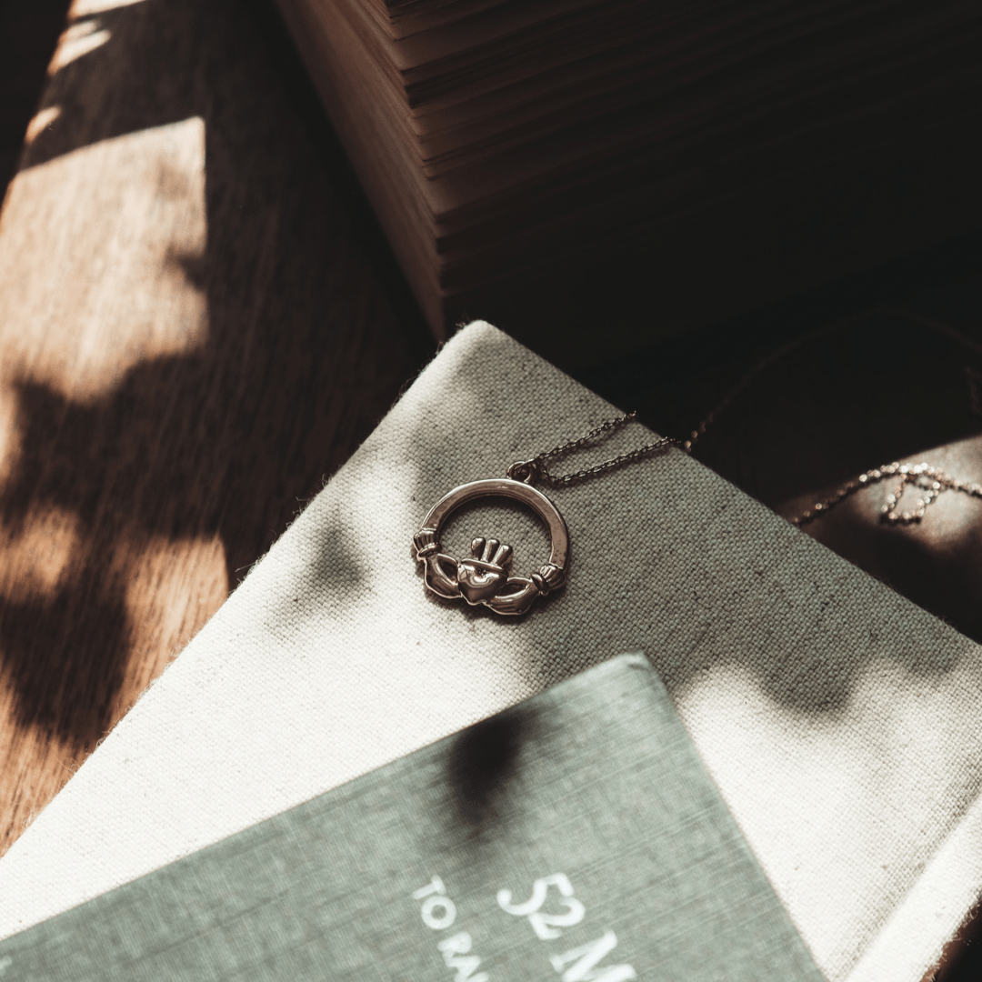 The Claddagh Necklace on a stack of books in dappled sunlight.