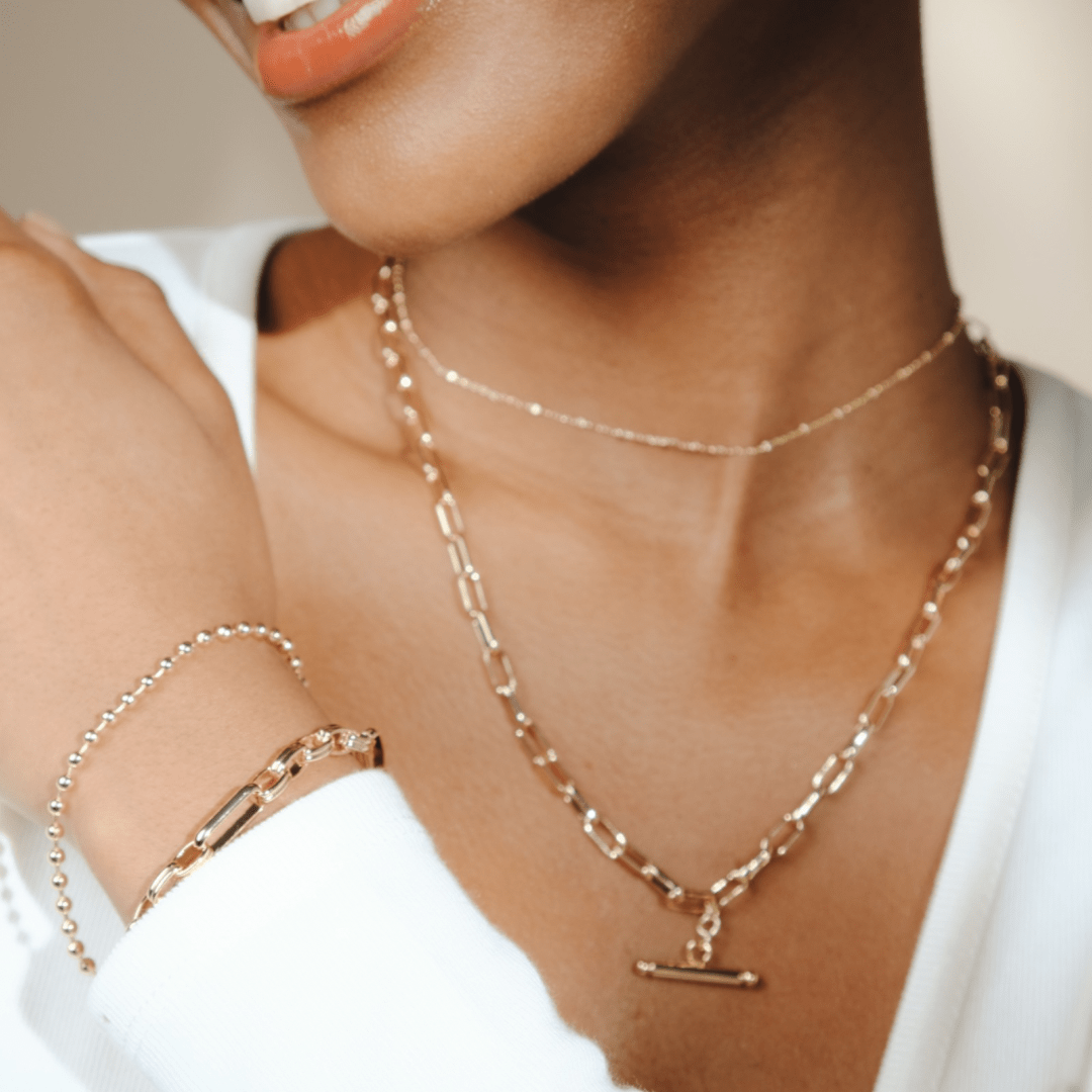 Close-up of a woman's neck and wrist, adorned with two gold necklaces and a T-Bar Link Chain bracelet.