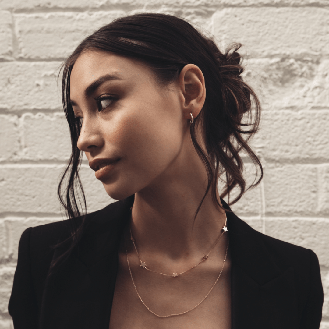 A woman wearing a blazer and delicate necklaces, including The Box Multi Star, poses against a white brick wall.