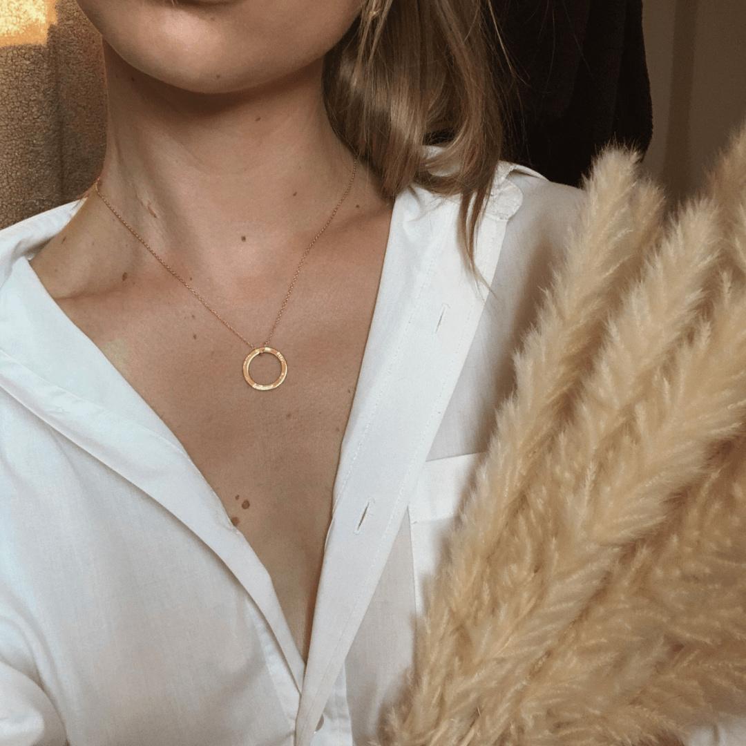 A person wearing a white shirt and a gold circular pendant necklace is holding Dá fhada an lá tagann an tráthnóna.