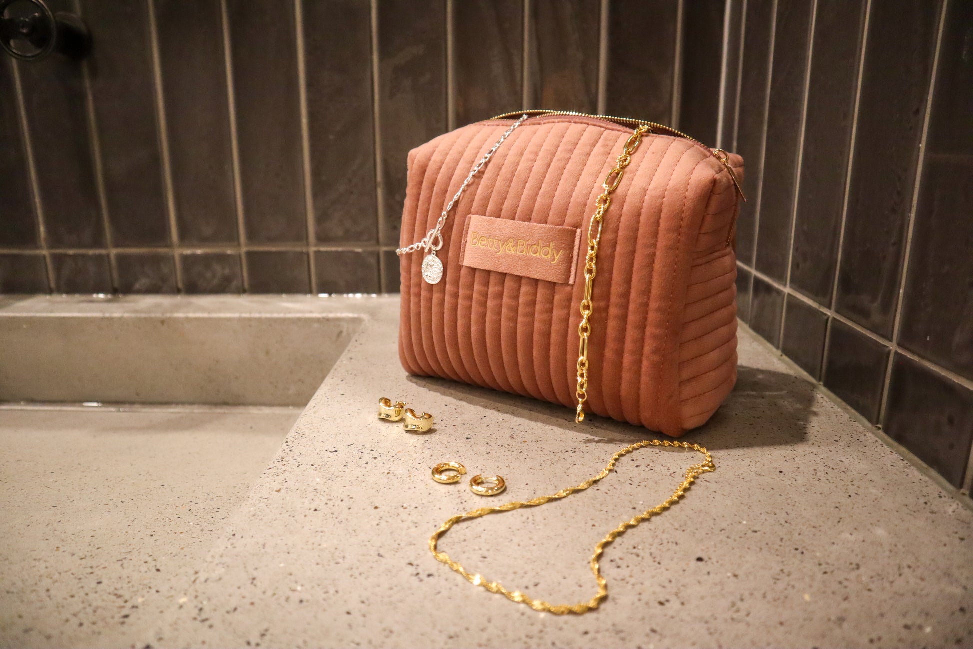 A pink quilted Betty & Biddy Jewellery Travel Bag sits on a stone surface with scattered gold and silver jewelry.