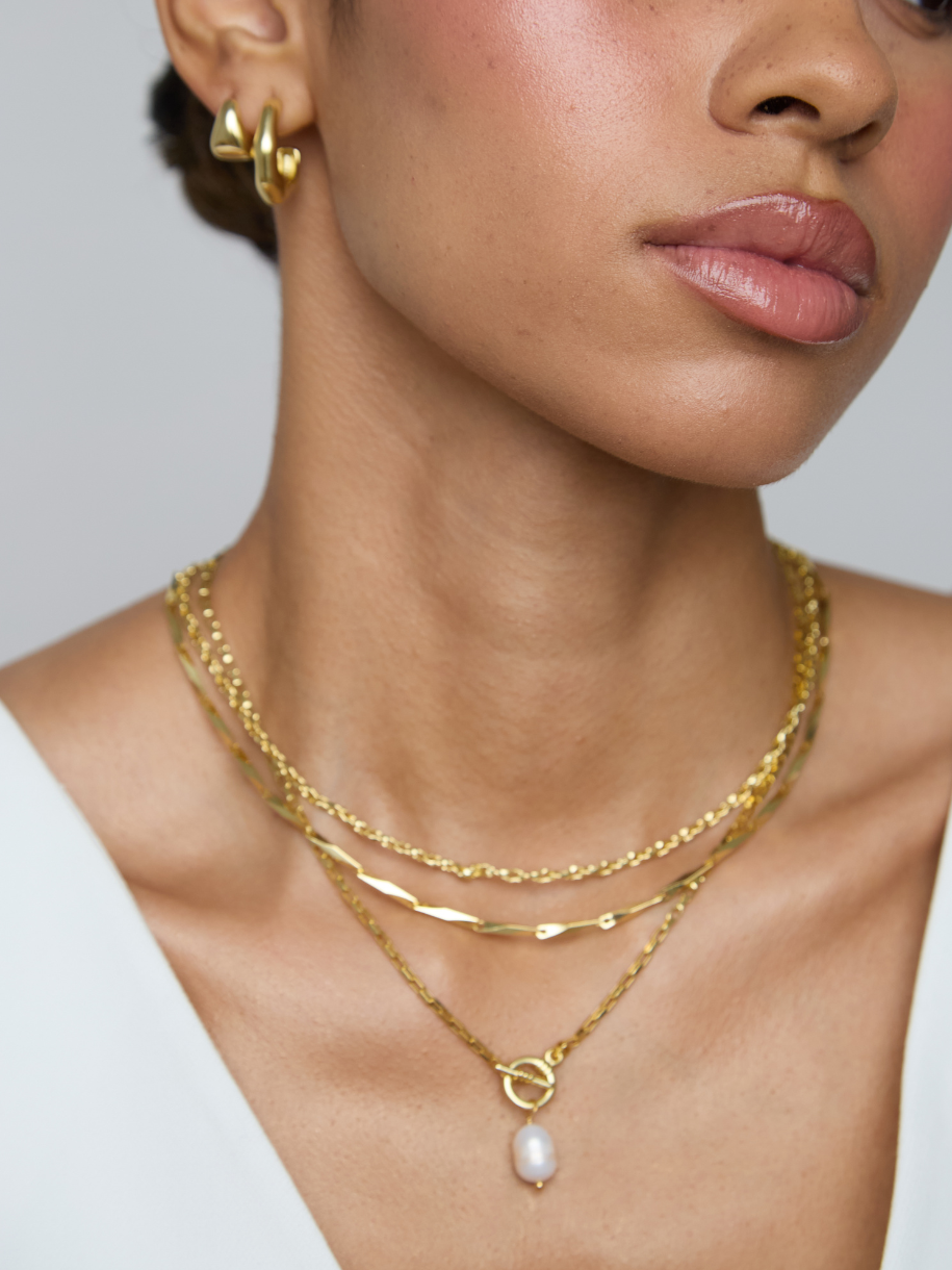 Close-up of a model wearing layered gold necklaces, including a delicate chain, a textured chain, and a pendant necklace with a white pearl drop, along with matching gold hoop earrings.