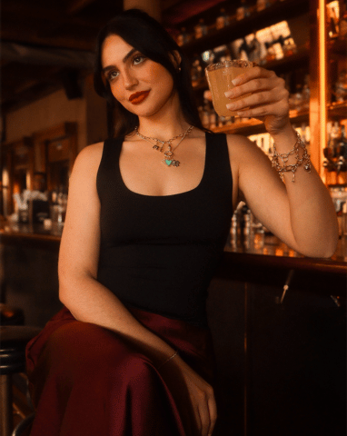 A woman with dark hair and red lipstick holds a drink while sitting at a bar.