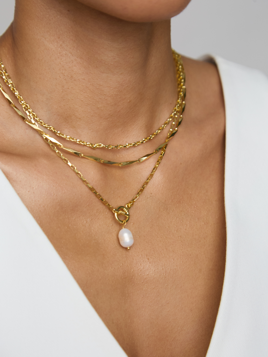 A close-up of a model's neck and chest wearing a layered gold necklace with a single white pearl pendant, paired with a white V-neck top.