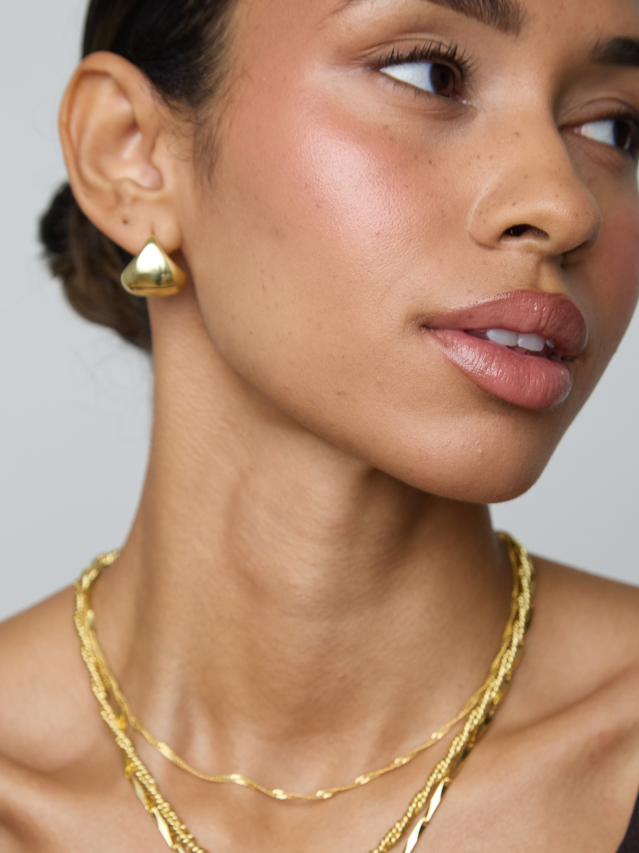 Close-up of a woman wearing a gold geometric stud earring and layered gold necklaces against a plain light gray background.
