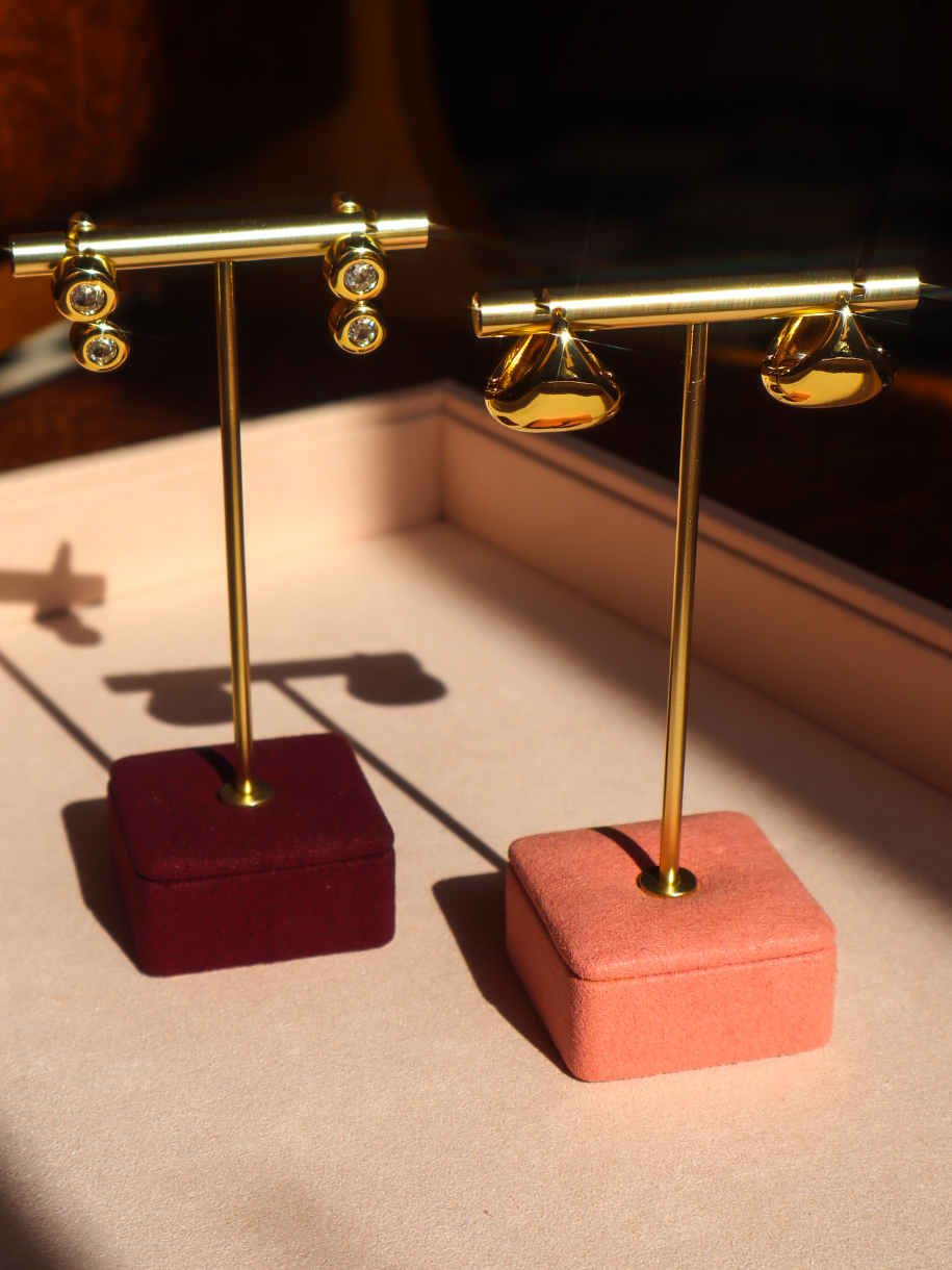 Two pairs of gold-toned stud earrings displayed on gold T-stands with velvet jewelry boxes, set on a beige surface with warm directional sunlight and shadows.
