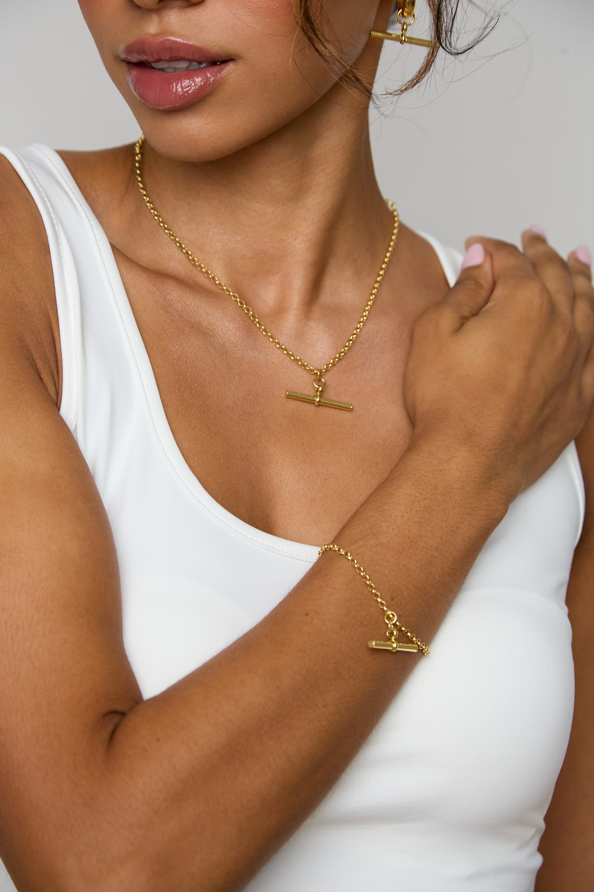 A woman wearing gold jewelry, including a gold necklace with a horizontal bar pendant, matching drop earrings, and a gold ring, paired with a white tank top against a light gray background.