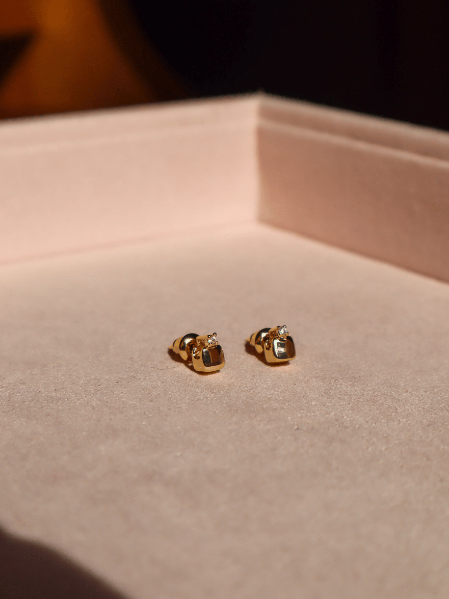 A pair of small gold stud earrings resting on a beige jewelry display box.