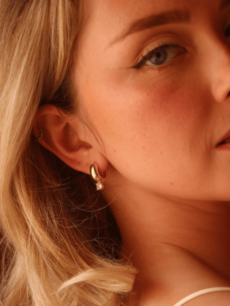 A close-up lifestyle shot of a woman with blonde hair wearing a small gold hoop earring with a tiny clear crystal accent.