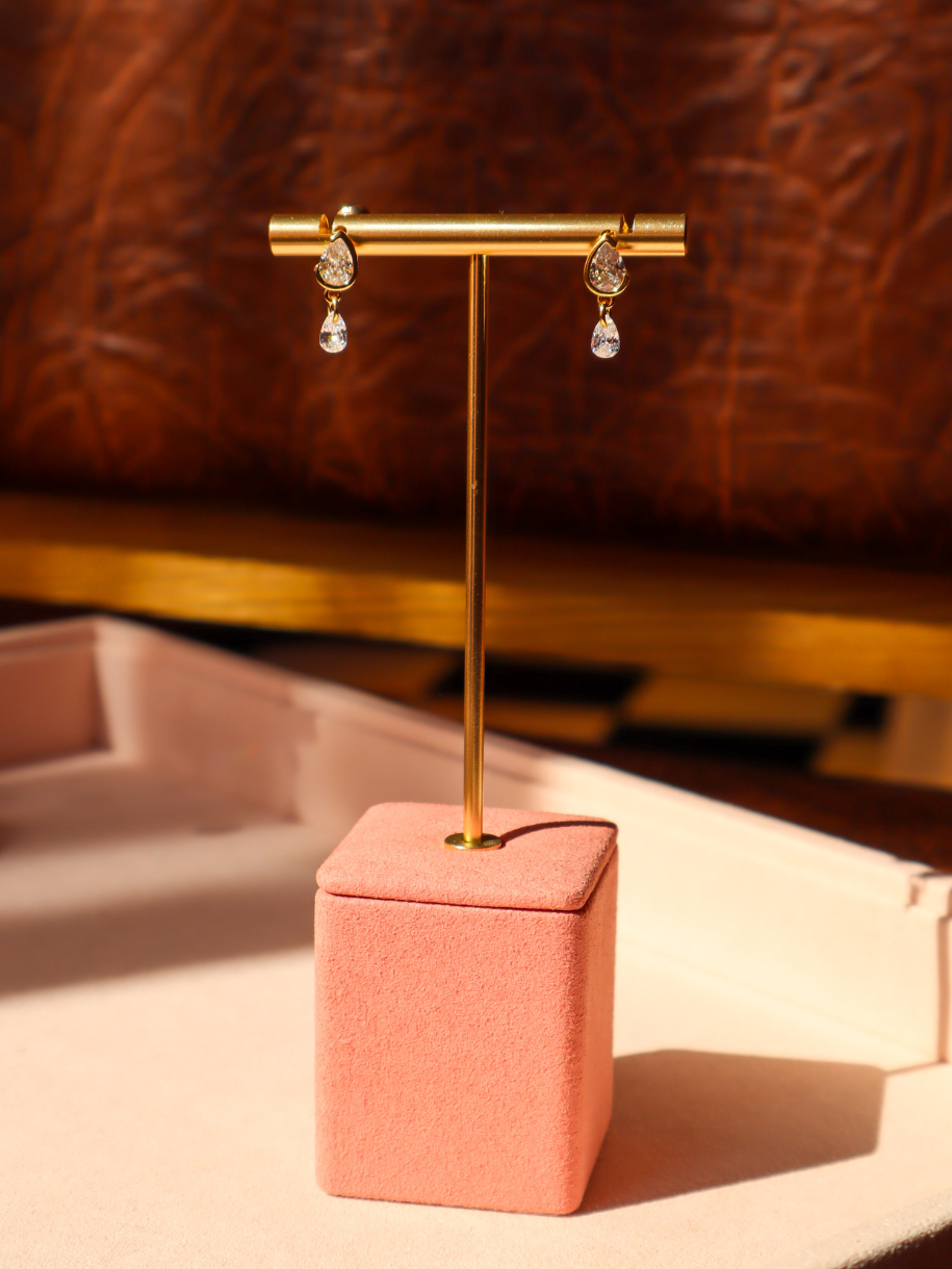 A pair of dangling earrings displayed on a gold T-stand with a pink velvet jewelry box base, set against a warm brown leather background.