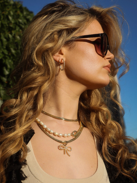 A woman with wavy blonde hair wearing a Pearl Bow Charm and sunglasses is seen in profile against a blue sky.