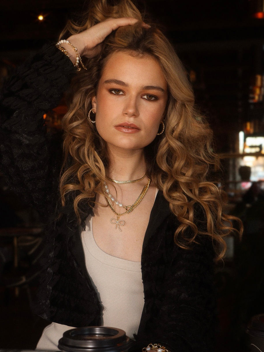 A woman with long, wavy blonde hair wearing Pearl Bow Charm jewelry poses in a dimly lit indoor setting.