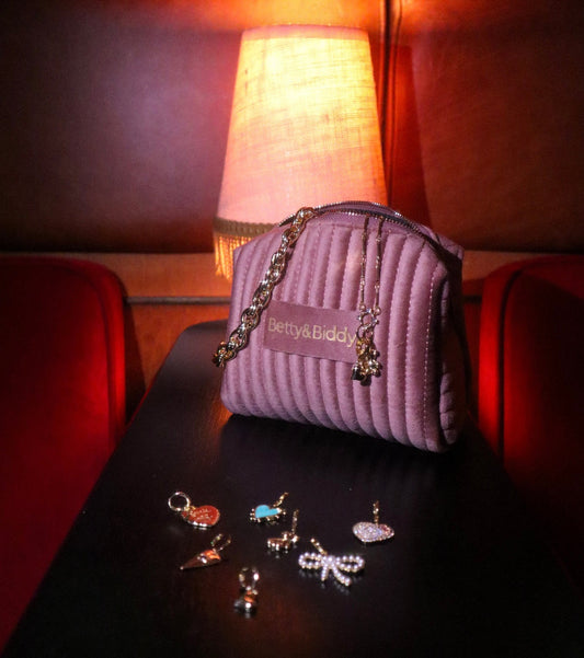 A velvet Jewellery Travel Bag and jewelry charms sit on a table under the light of a lamp.