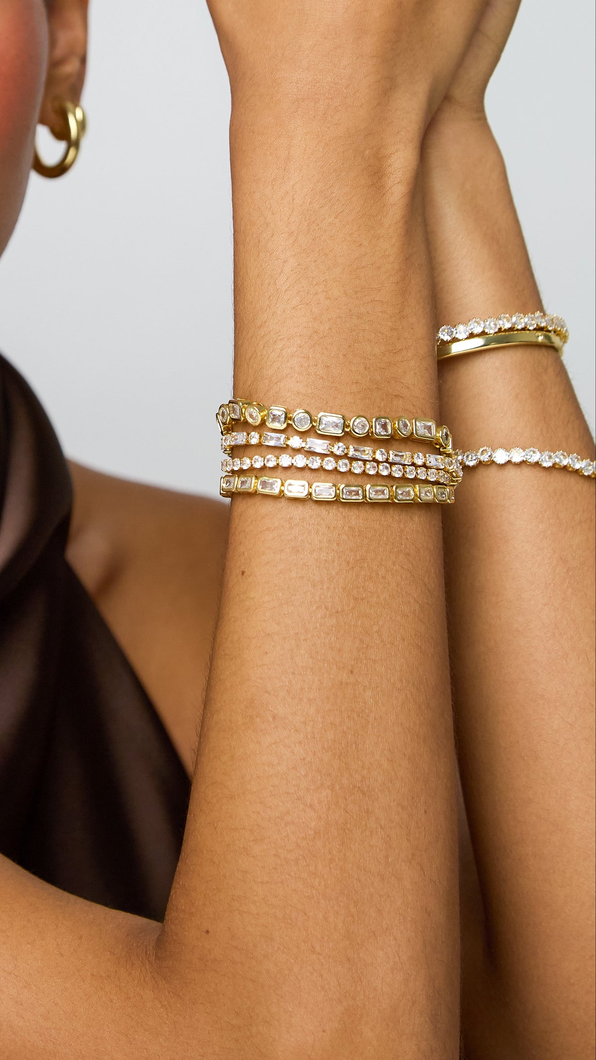 A close-up of a woman wearing gold jewelry, including a gold hoop earring and stacked gold bracelets with clear stones, against a light gray background.