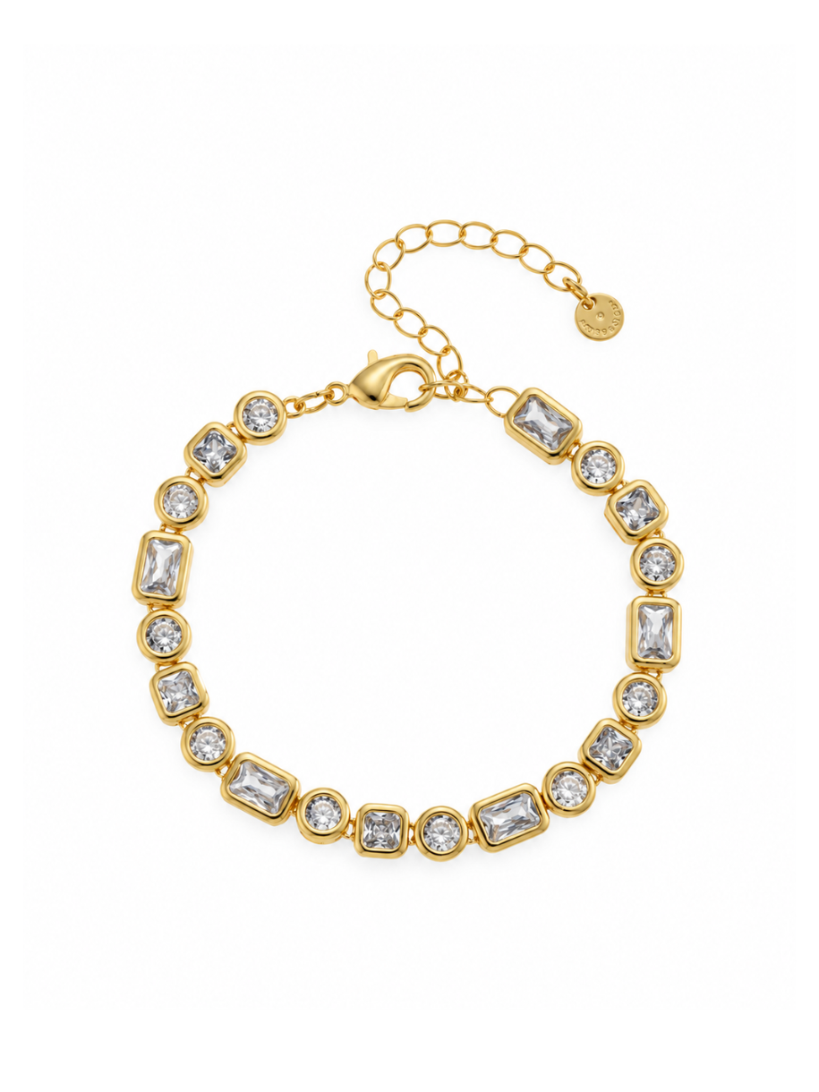 The Mixed Cut Bezel Tennis Bracelet features an alternating row of emerald-cut and round clear stones, each set in a polished gold bezel. A gold chain extension and logo charm hang from the lobster clasp