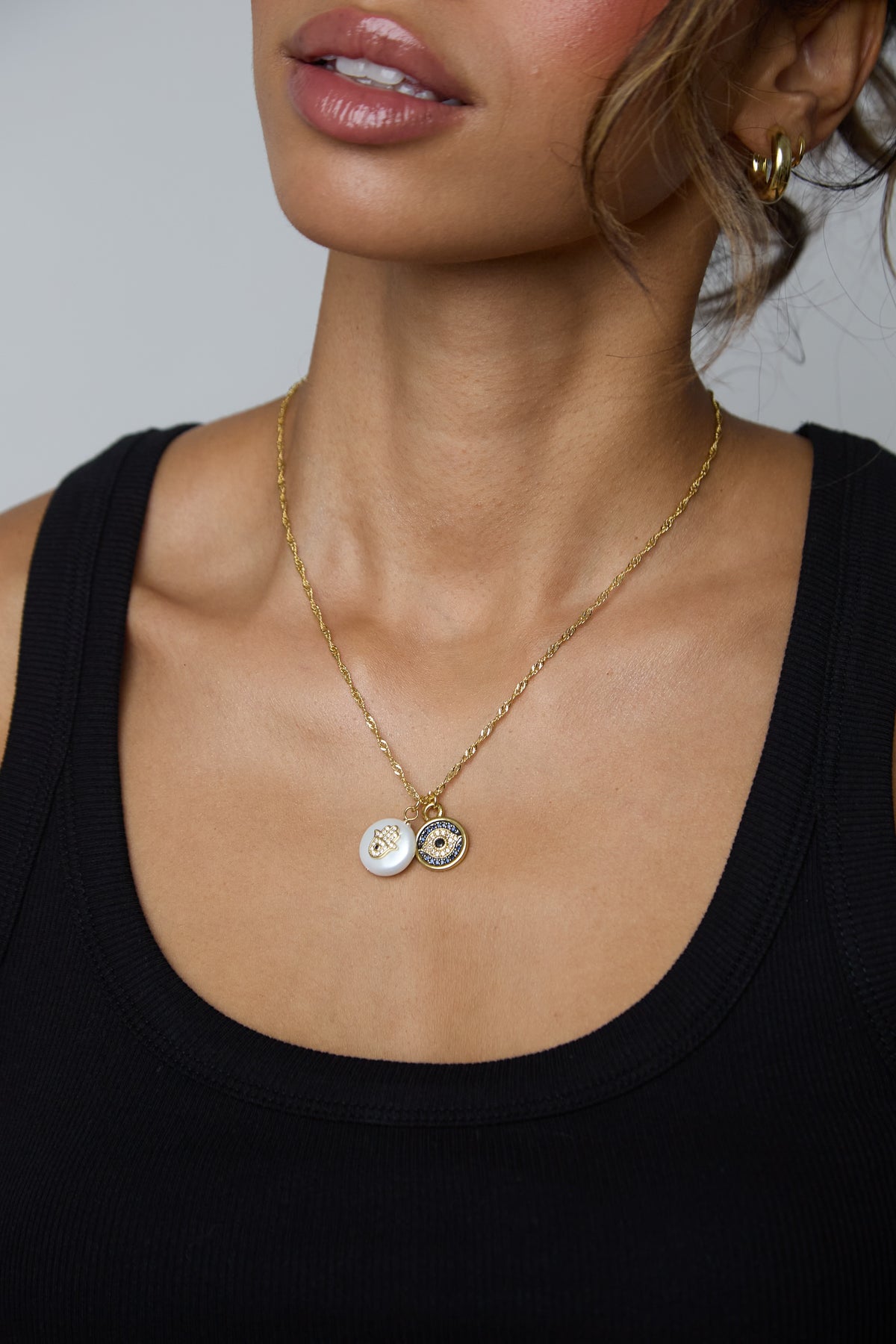 A close-up of a model wearing a delicate gold chain necklace with two round white and black enamel pendants, styled with a black ribbed tank top.