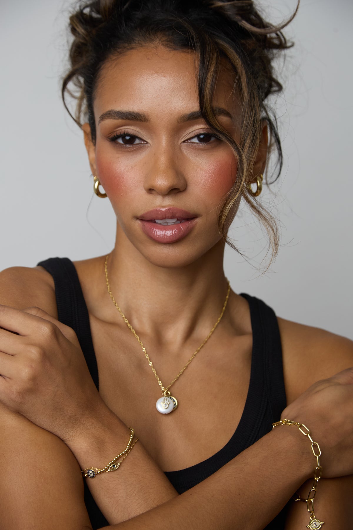 A close-up portrait of a woman wearing gold jewelry, including a delicate gold chain necklace with a round white pendant, a gold hoop earring, and a gold bracelet, against a plain light gray background.