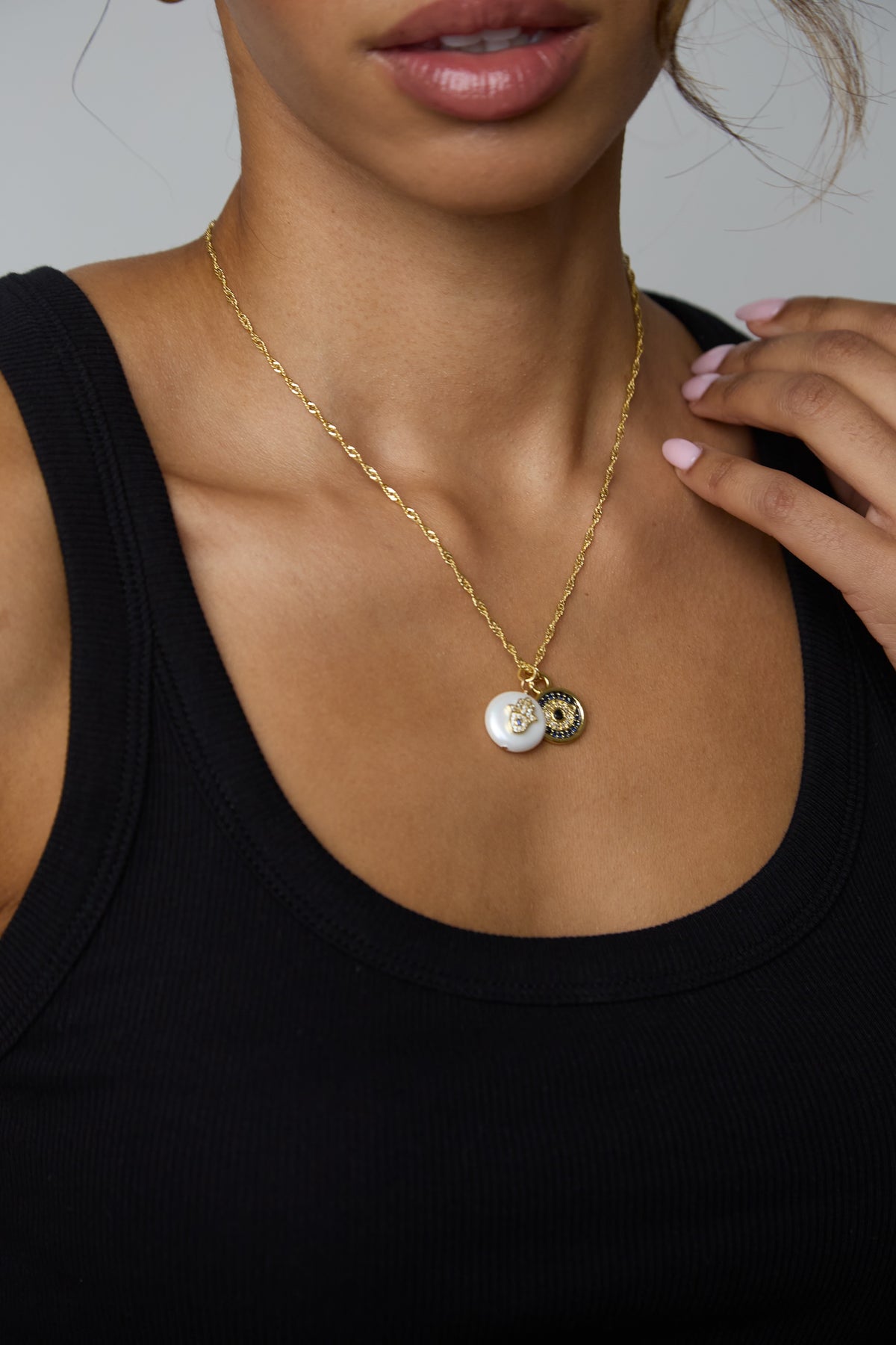 A close-up of a model wearing a delicate gold chain necklace with two small circular pendants, one white and one gold, against a black ribbed tank top.