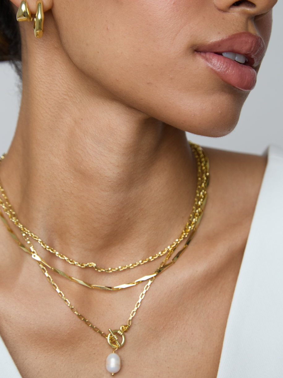 Close-up of a model wearing a layered gold necklace with a single white pearl pendant, along with a gold hoop earring and white top.