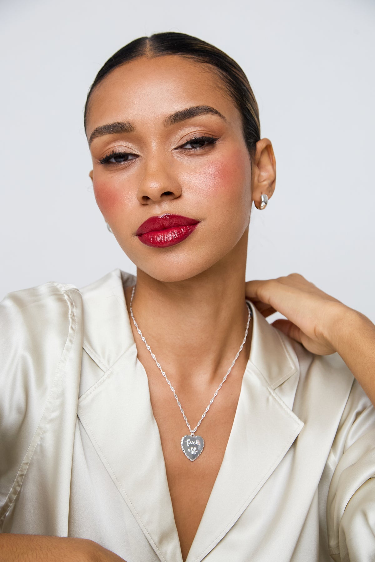 A woman with her hair pulled back wears a silver heart-shaped pendant necklace with a clear central stone, paired with a cream satin blazer against a light gray studio background.