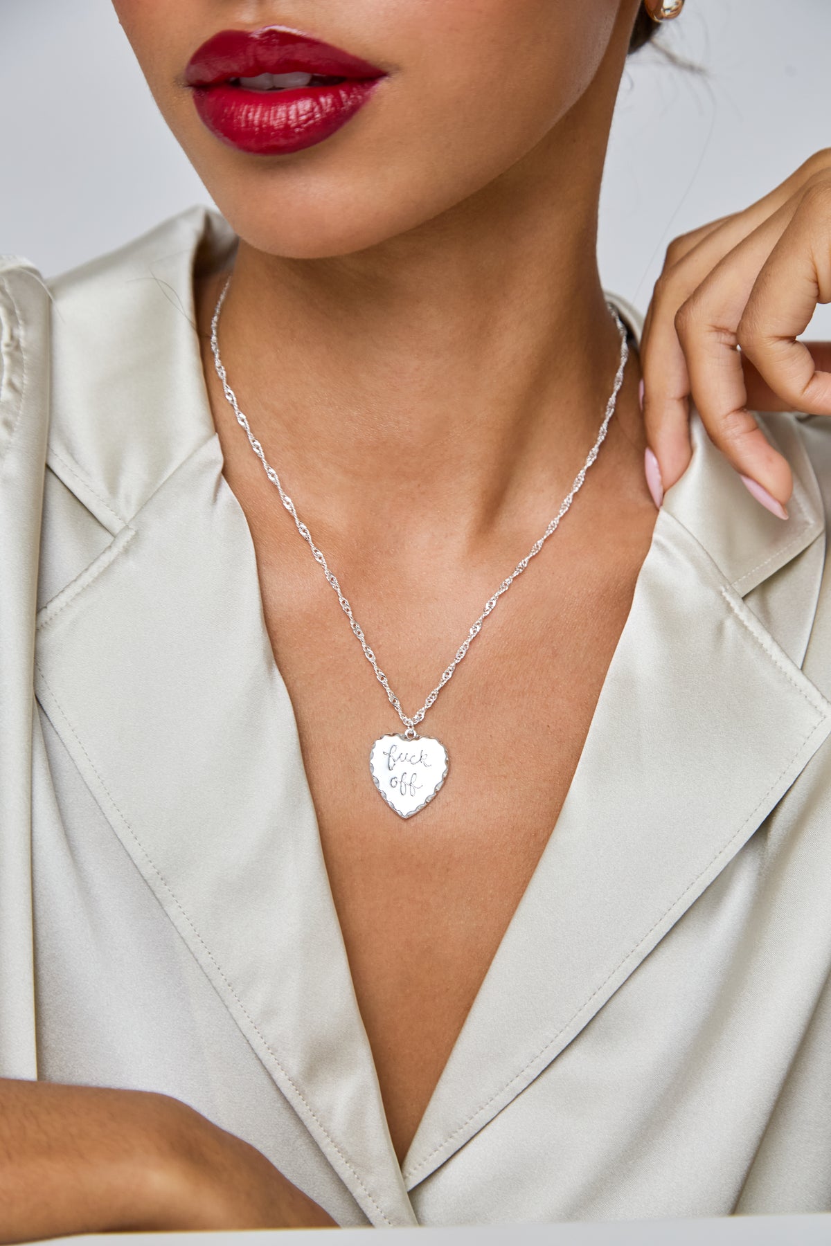 A close-up of a model wearing a silver heart-shaped pendant necklace engraved with 'Love You' on a delicate chain, styled with a cream satin blazer.