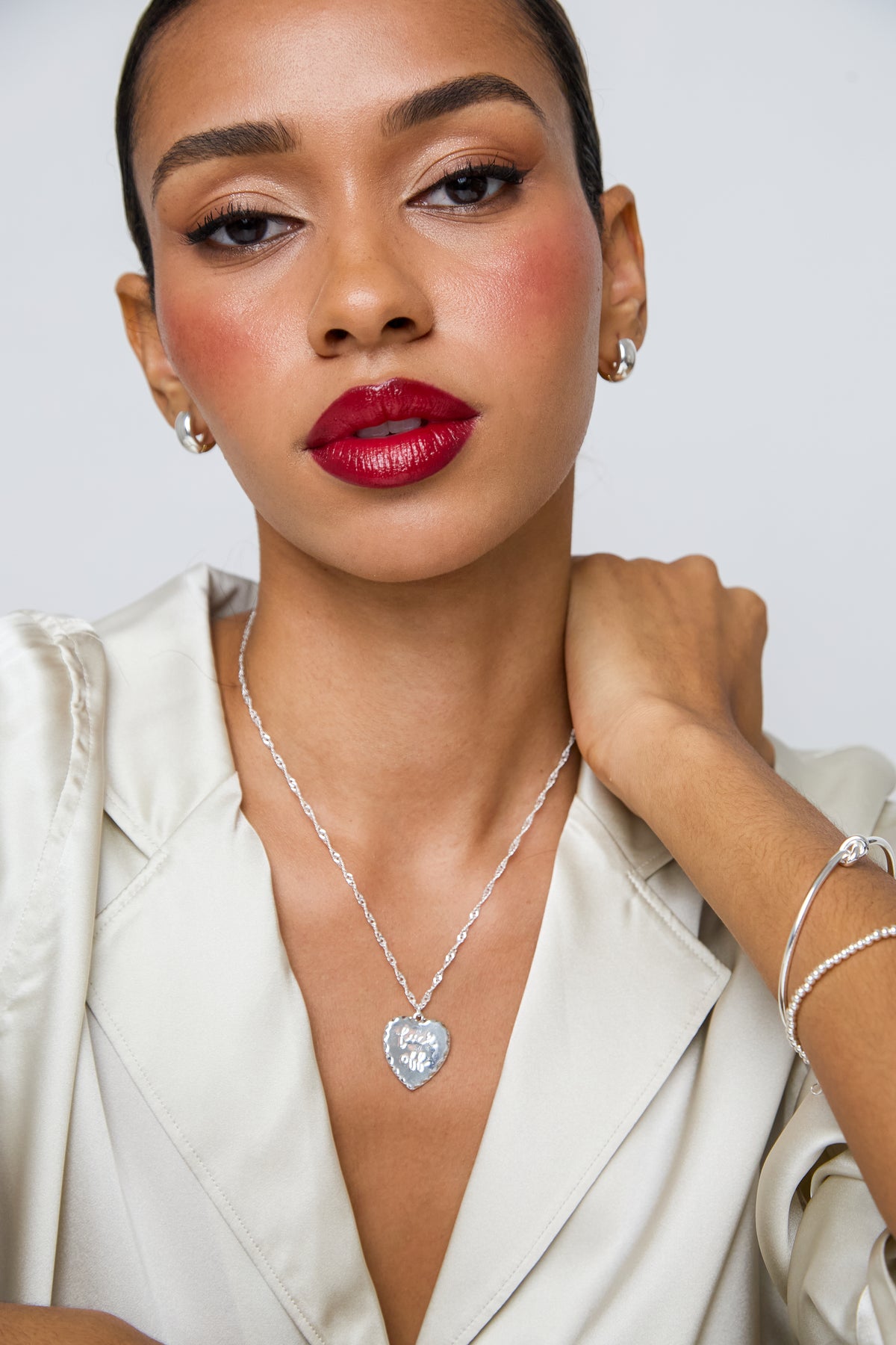 A close-up of a woman wearing a silver heart-shaped pendant necklace with a clear center stone, styled with a cream satin blouse against a light gray background.