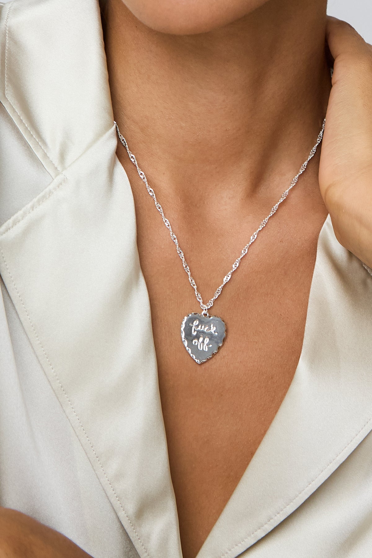 A close-up of a model wearing a silver heart-shaped pendant necklace with the engraved text 'fuck off' in black script, styled with a cream satin blazer.