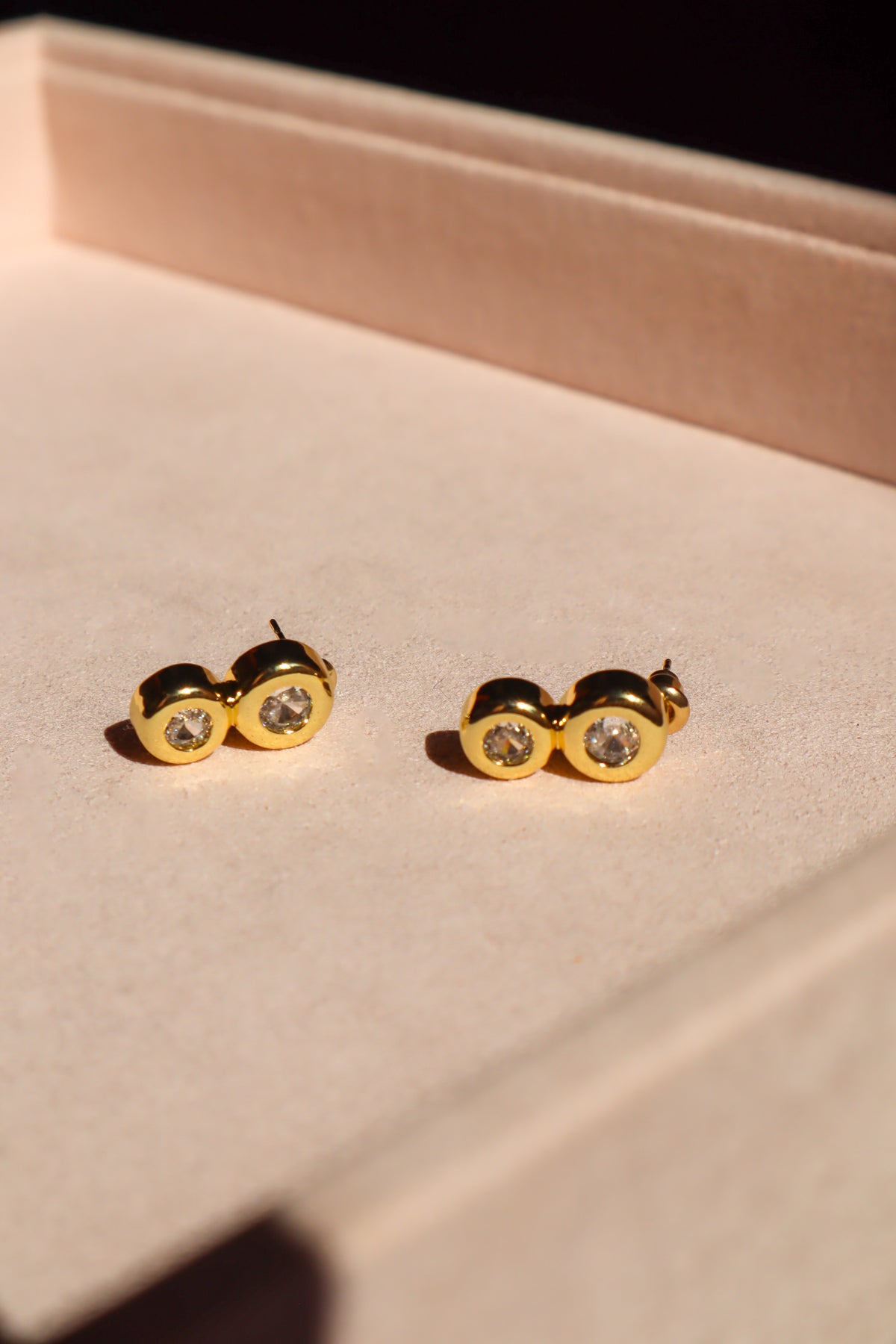 A pair of gold stud earrings with clear round gemstones, resting on a beige textured jewelry box.