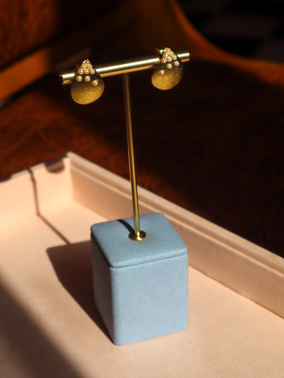 A pair of textured gold dangle earrings with small pearl-like accents, displayed on a gold stand with a light blue velvet base on a beige tray.